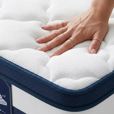 A close-up of a white quilted mattress topper or mattress surface being pressed down by a person's hand, with a navy fabric border and part of a product tag visible.