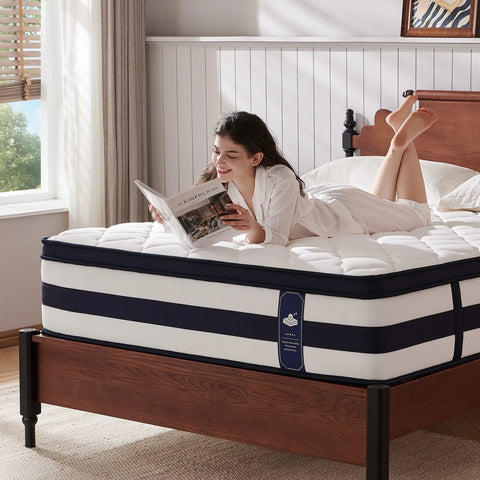 A woman in a white robe lies on a striped mattress on a wooden bed frame, reading a magazine in a bright bedroom. The mattress has white and navy blue horizontal stripes with a small blue brand tag on the side.