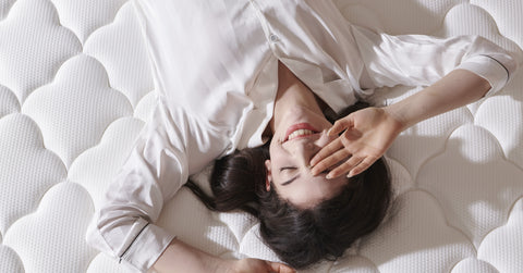 Woman lying on a white mattress with a smile, wearing a white shirt.